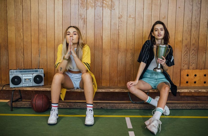 Two young women sitting in a gym, one holding a trophy, with a vintage boombox beside them.