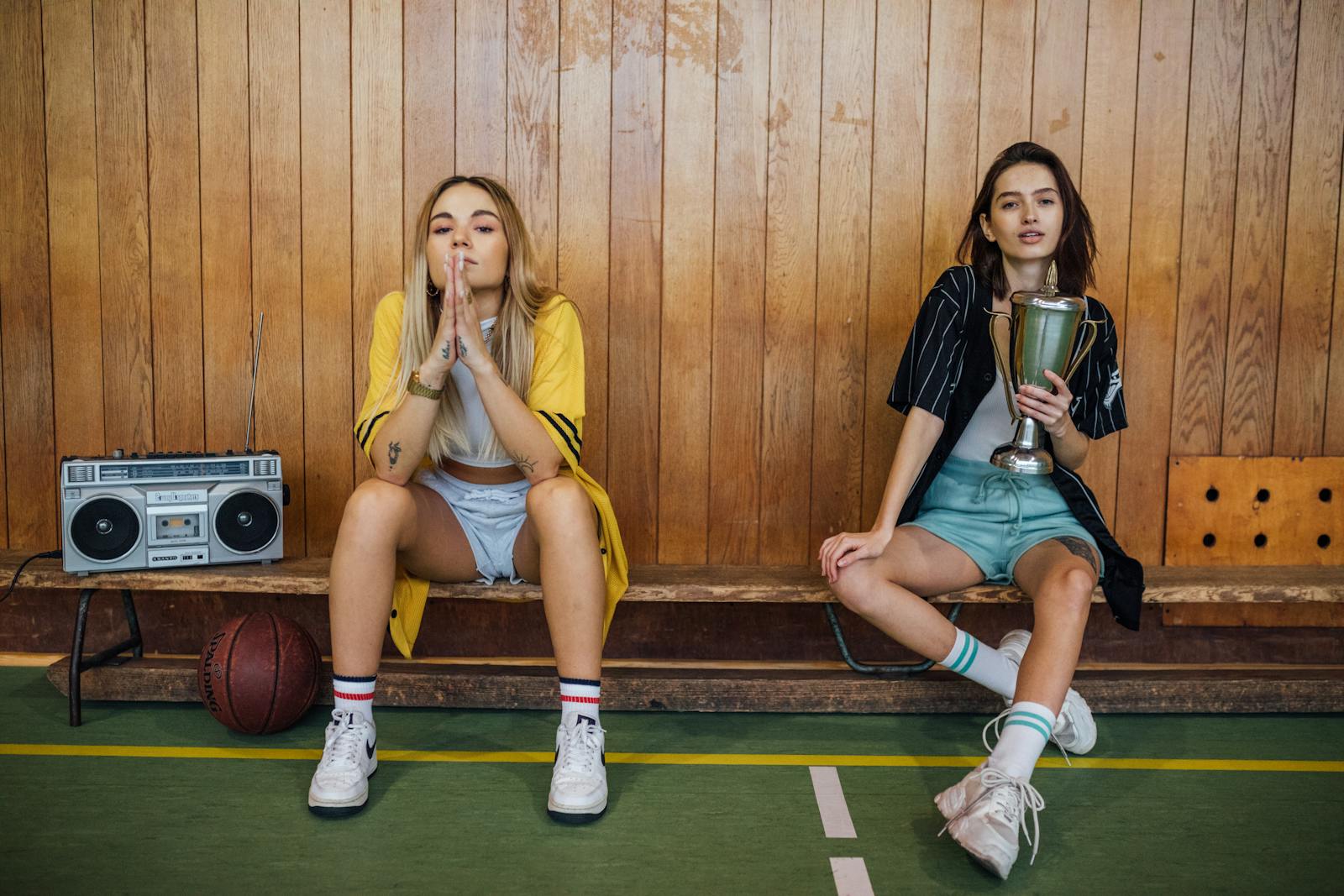 Two young women sitting in a gym, one holding a trophy, with a vintage boombox beside them.