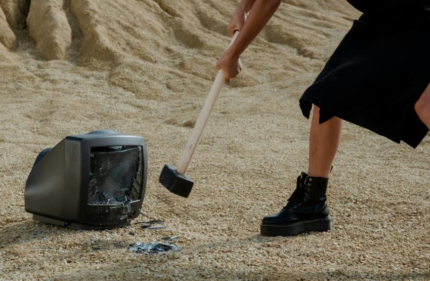 A person in black boots and dress smashes an old television with a mallet on sandy ground.