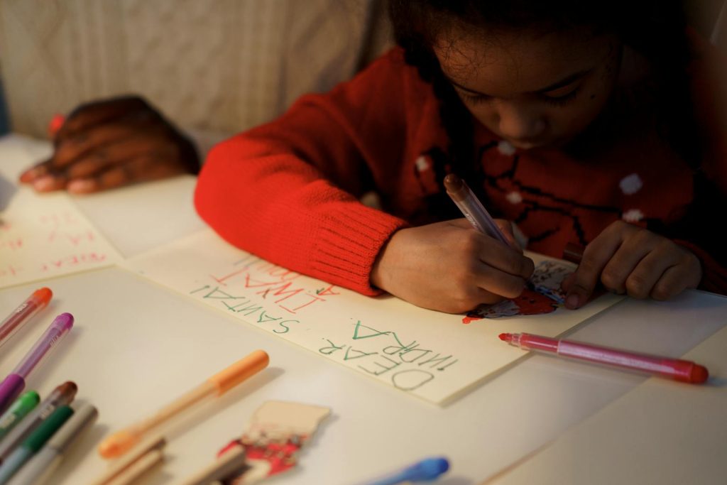 Young girl writing a letter to Santa Claus with colorful pens indoors, capturing holiday spirit.