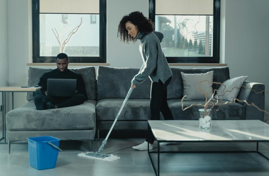 Man working on laptop while woman mops floor in stylish living room.