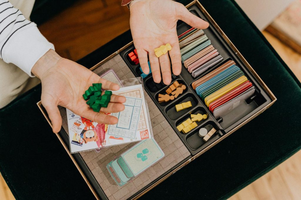 Colorful board game components being held in hands above a game box, top view.