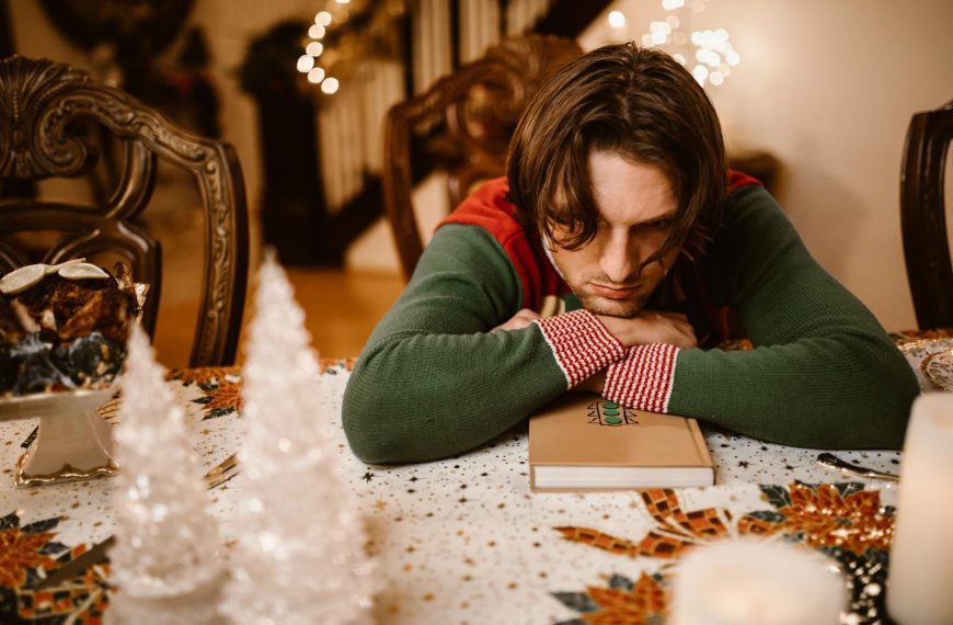 A man in a sweater looks thoughtful and sad at a decorated Christmas table indoors.
