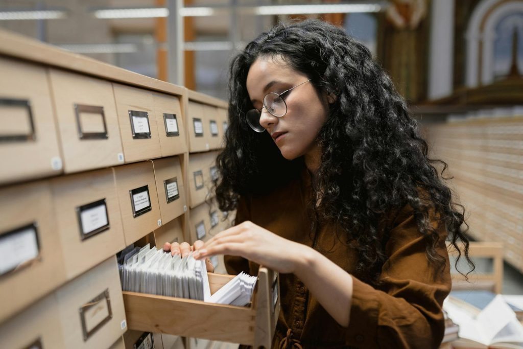 Curly haired woman in brown browsing a library card catalog with focus and curiosity.