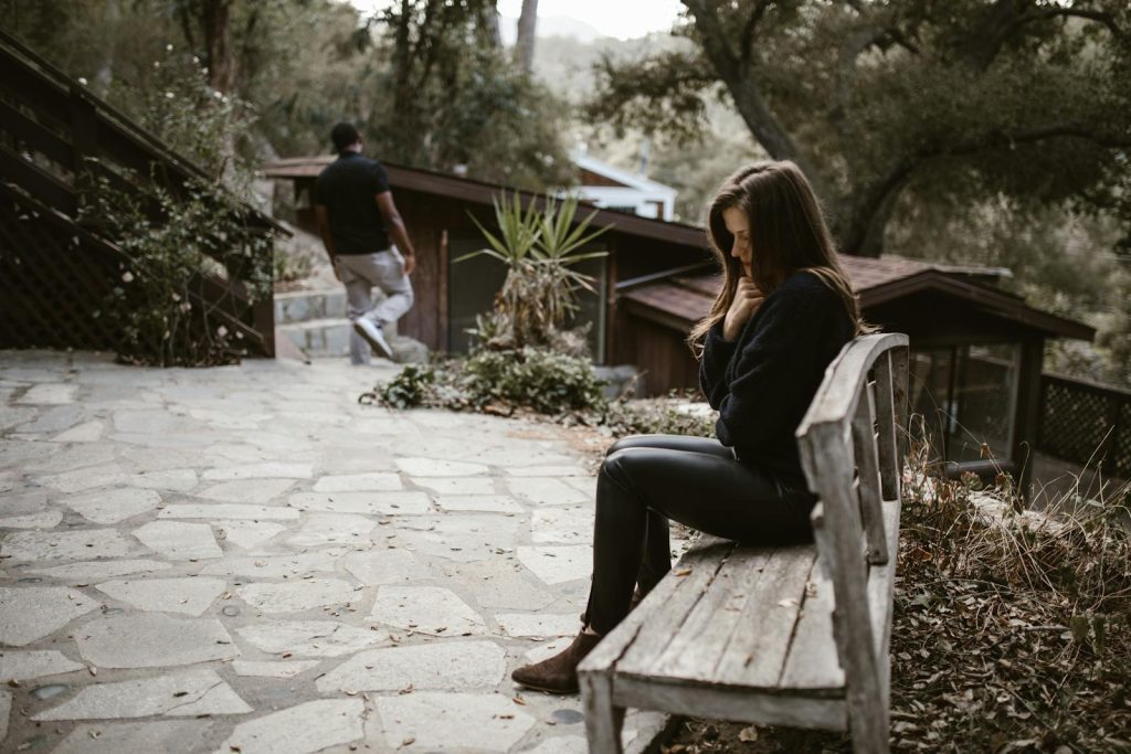 A woman sits pensively on a bench outside after a breakup, while a man walks away.