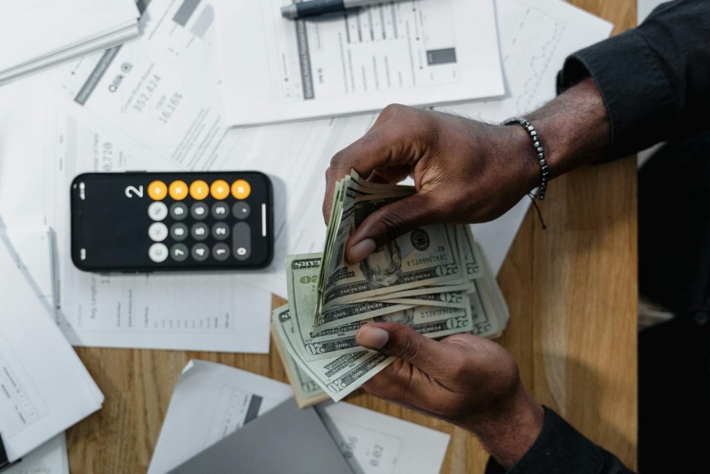 Person counting dollar bills over documents with a smartphone calculator on the desk.