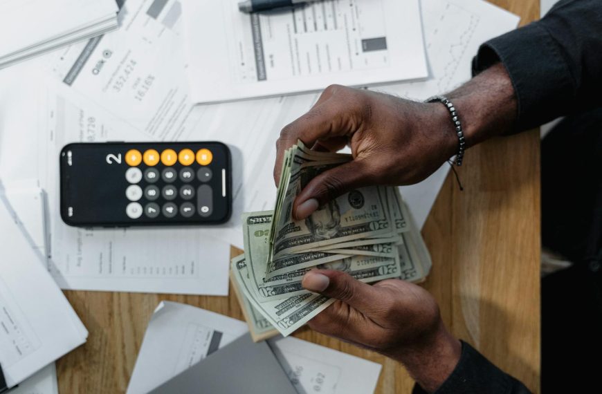 Person counting dollar bills over documents with a smartphone calculator on the desk.