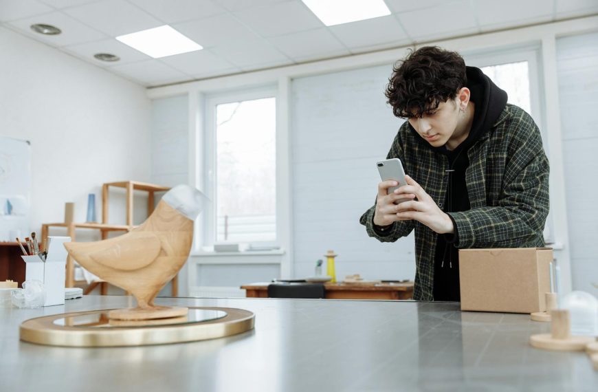 Young man captures a wooden sculpture using a smartphone in a well-lit indoor setting.
