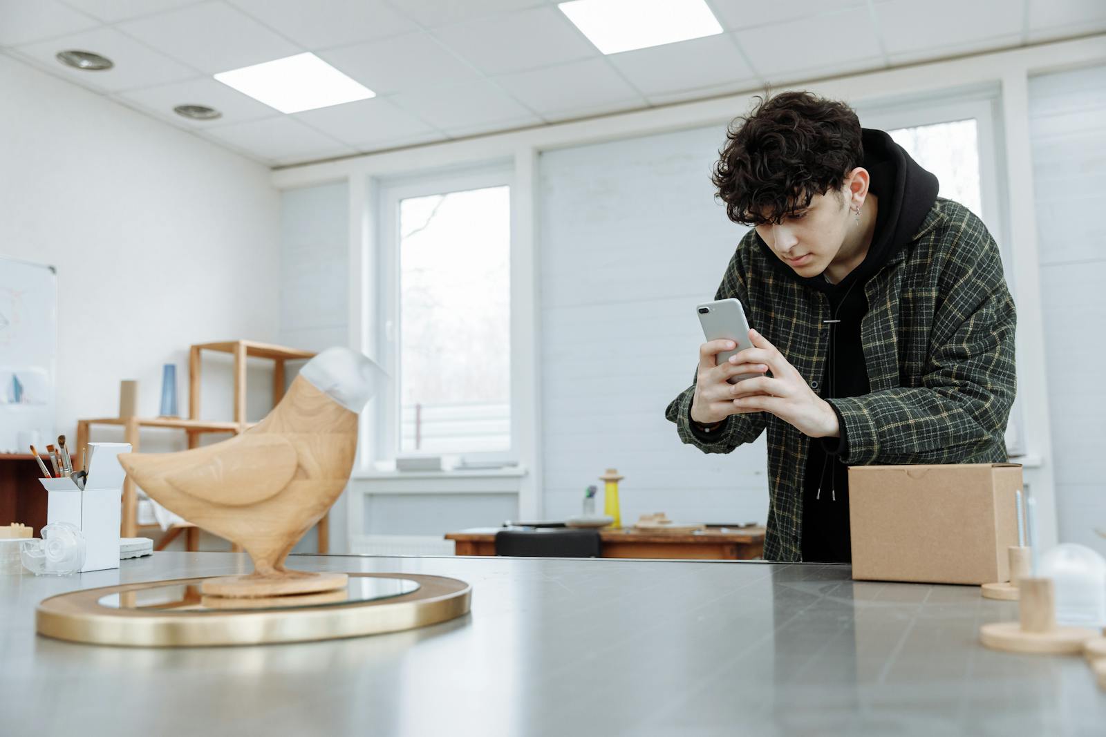 Young man captures a wooden sculpture using a smartphone in a well-lit indoor setting.