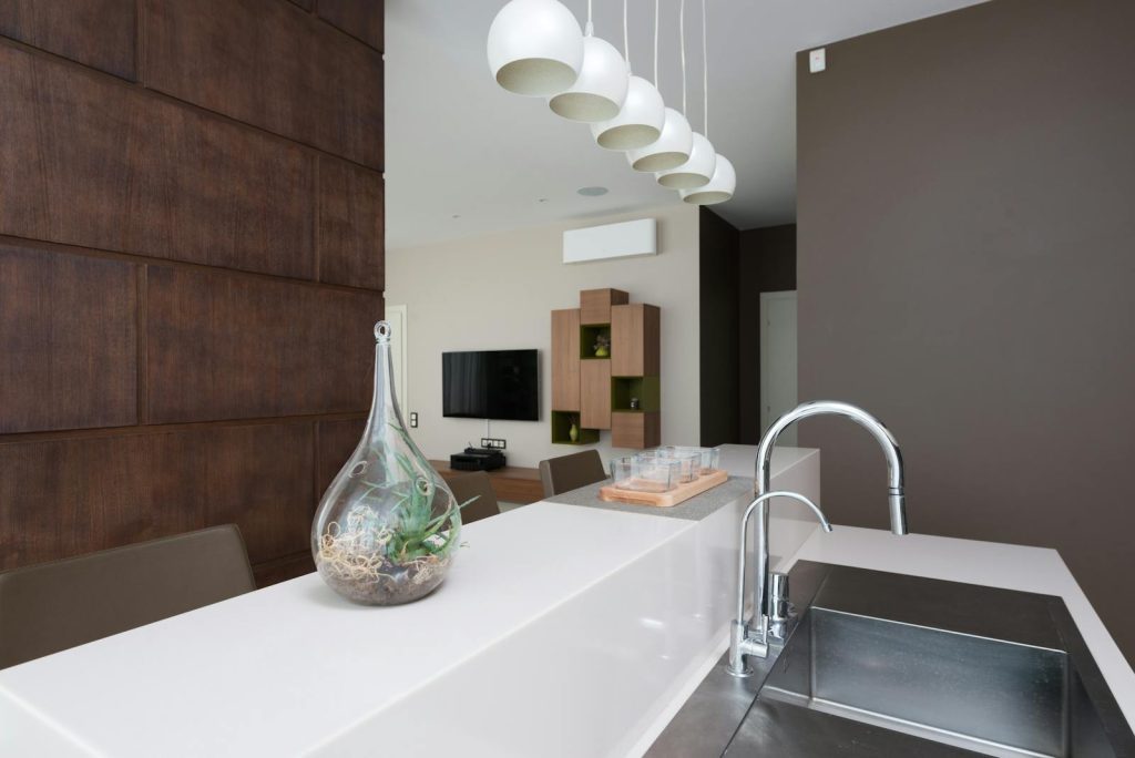 Decorative glass vase placed on white counter near sink with faucet in light modern apartment against decorated wall with TV
