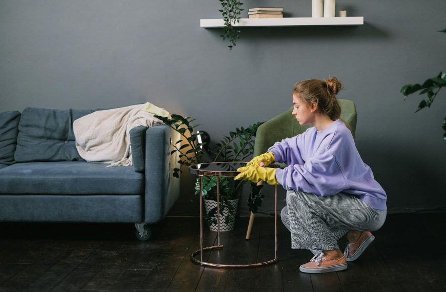 Side view of young female in latex gloves squatting down and wiping table in stylish living room in daytime