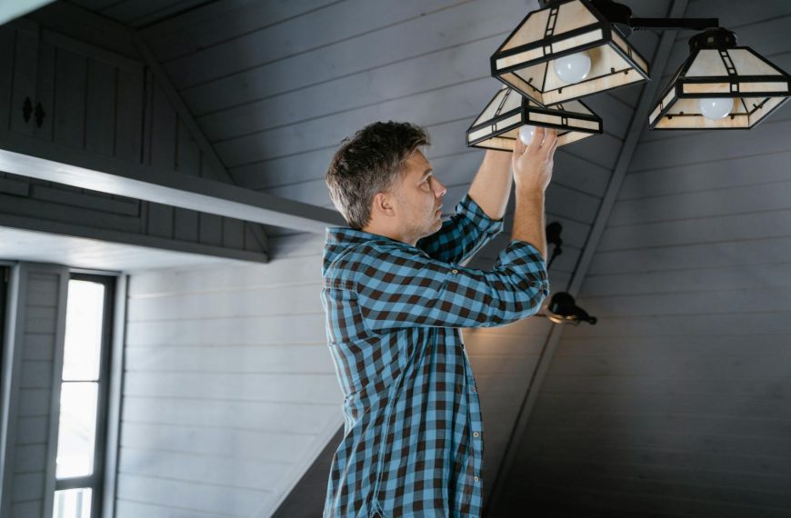 A man in a plaid shirt changes a lightbulb in a stylish attic room.