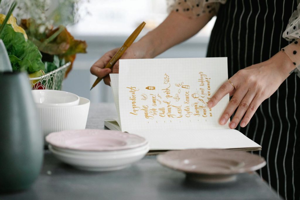 Woman writing a recipe in a notebook in a kitchen setting, surrounded by kitchenware.