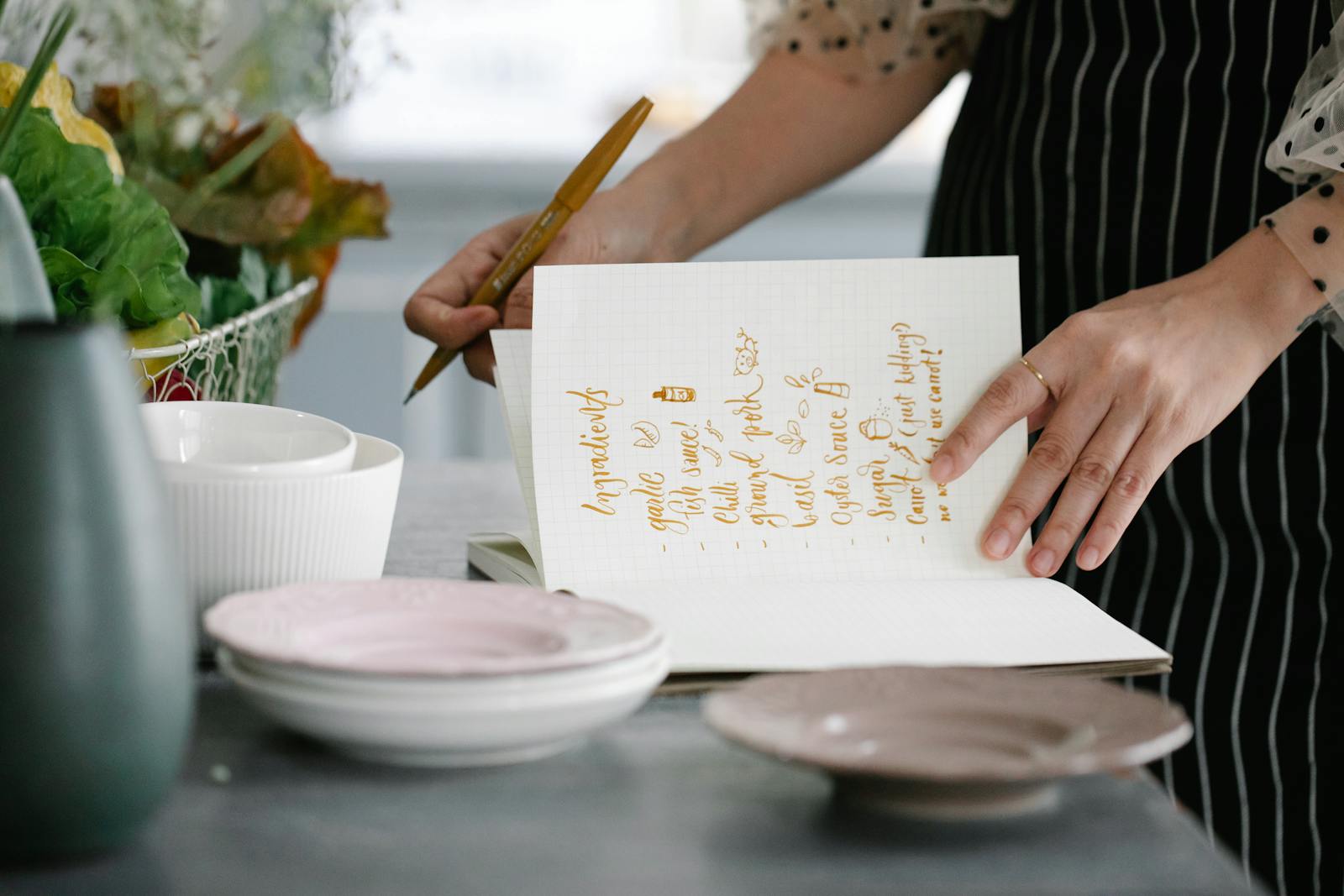 Woman writing a recipe in a notebook in a kitchen setting, surrounded by kitchenware.