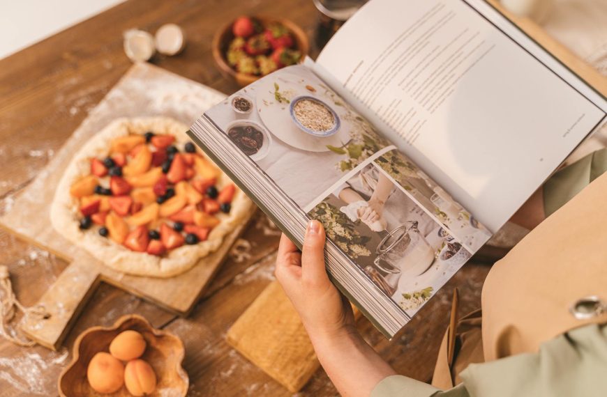 A person reads a cookbook while preparing a fruit tart on a wooden table.