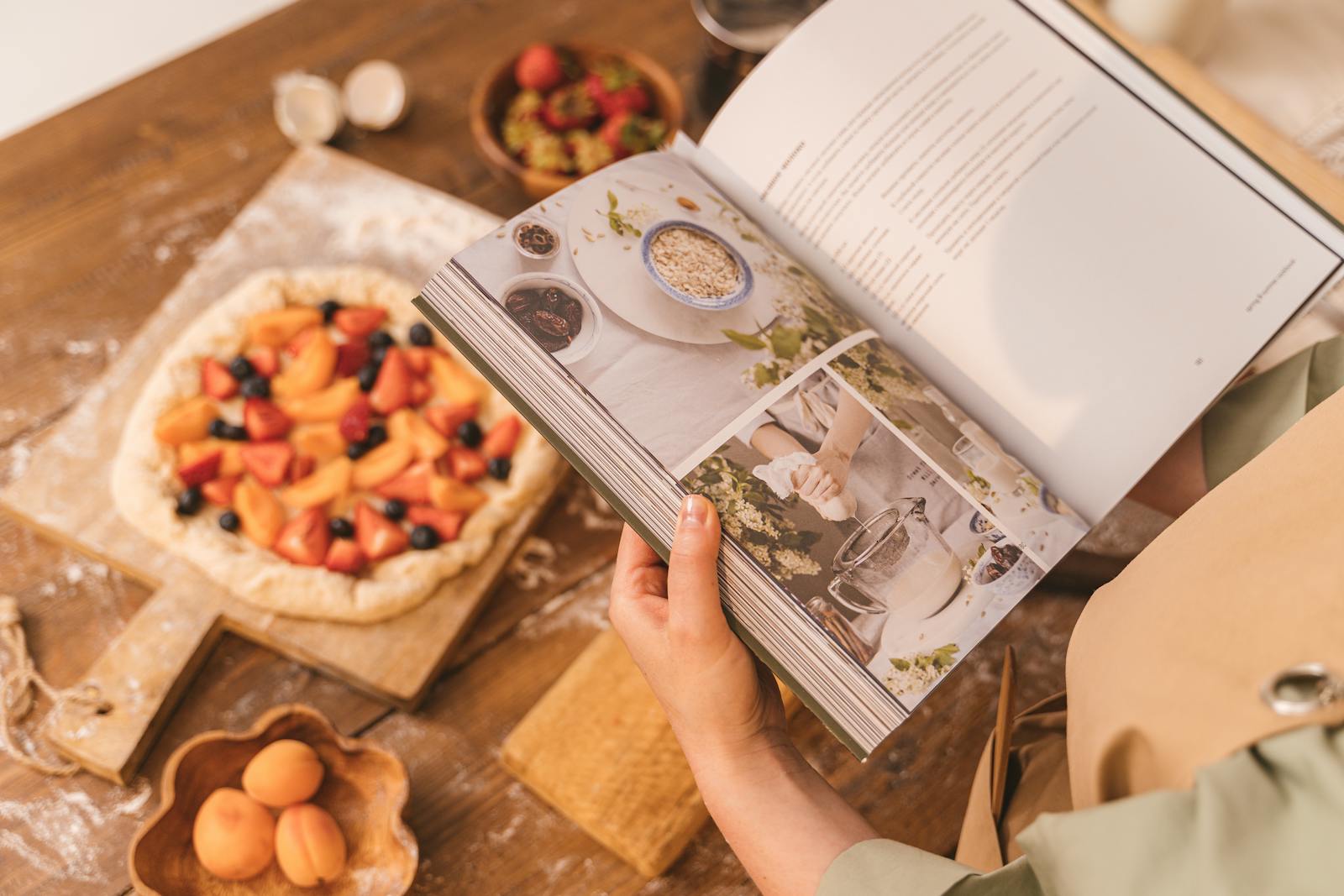 A person reads a cookbook while preparing a fruit tart on a wooden table.