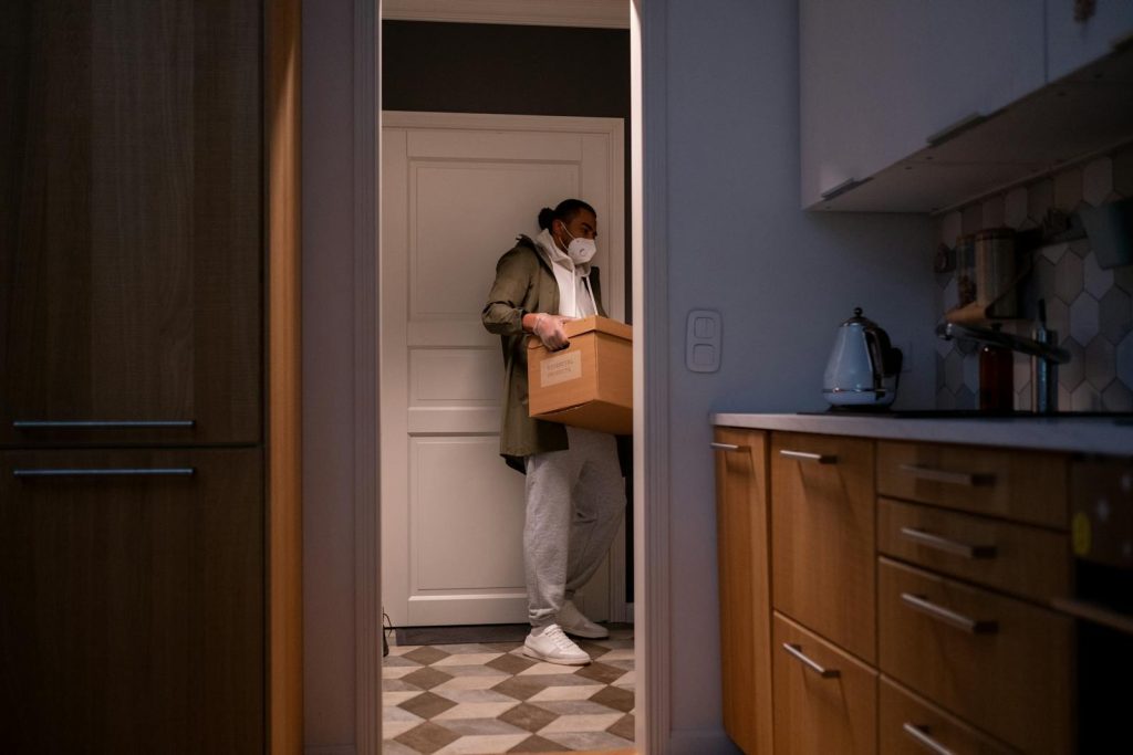 A man wearing a mask carries a box through a dimly lit kitchen, emphasizing safety and home logistics.