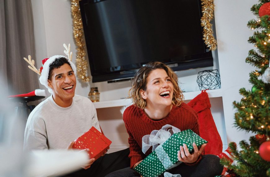 a person and a girl sitting on a couch with presents in front of a christmas tree