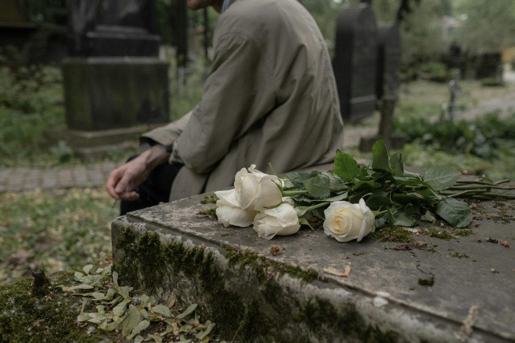 A person sitting by a tombstone with white roses, evoking themes of grief and remembrance.