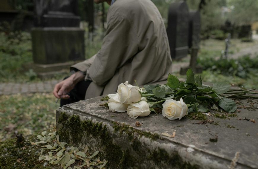 A person sitting by a tombstone with white roses, evoking themes of grief and remembrance.