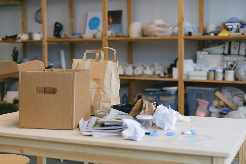 Cardboard box and paper bags on table in cluttered workspace with shelves. Ideal for packaging concepts.