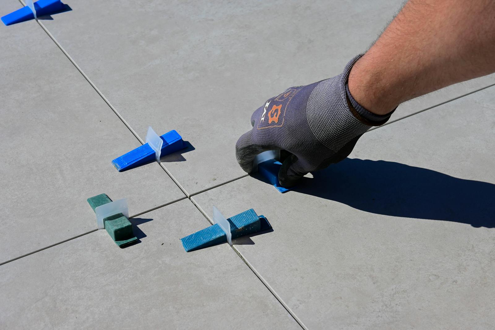 A close-up of a gloved hand installing tiles with spacers on a sidewalk.