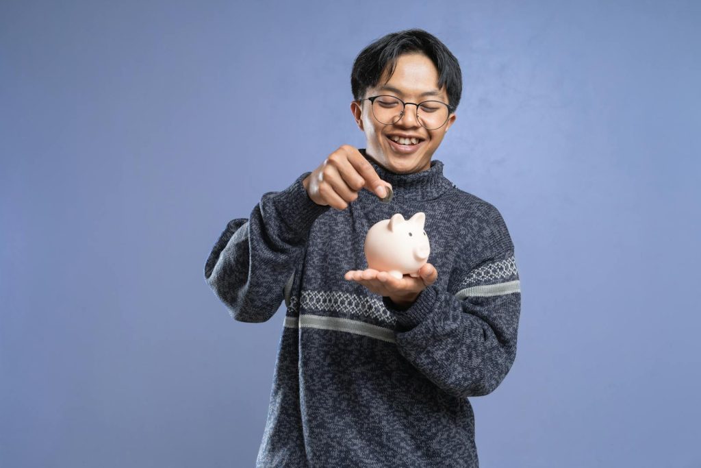 Smiling man holding a piggy bank and inserting a coin, representing savings and financial planning.