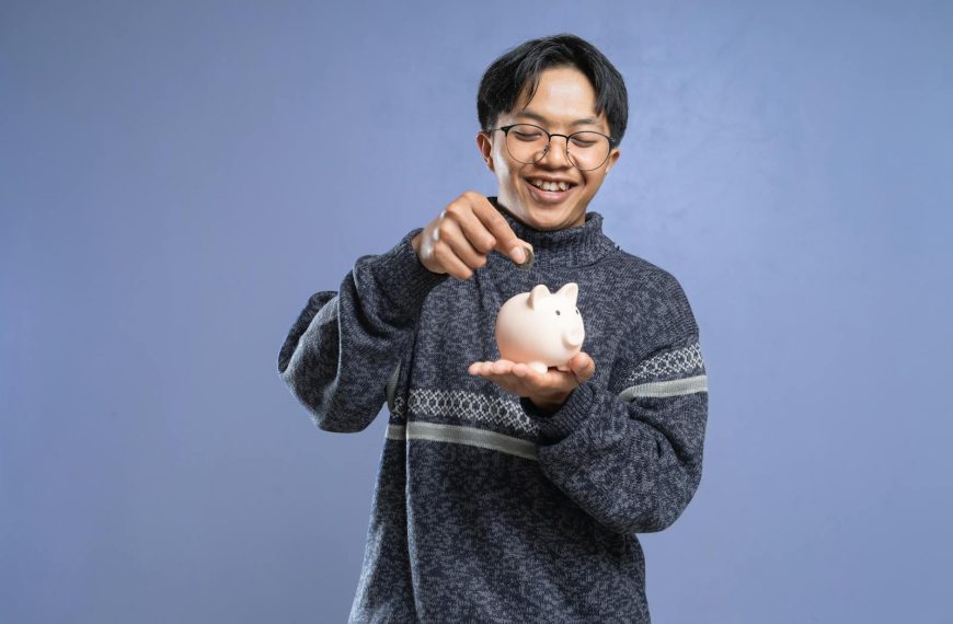 Smiling man holding a piggy bank and inserting a coin, representing savings and financial planning.