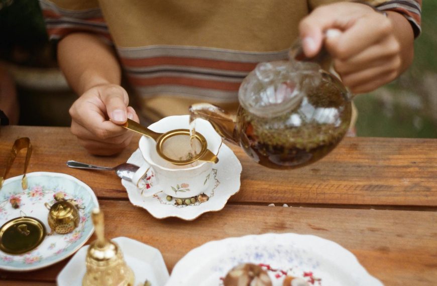 Close-up of a person pouring tea into a porcelain cup with floral design on a wooden table.