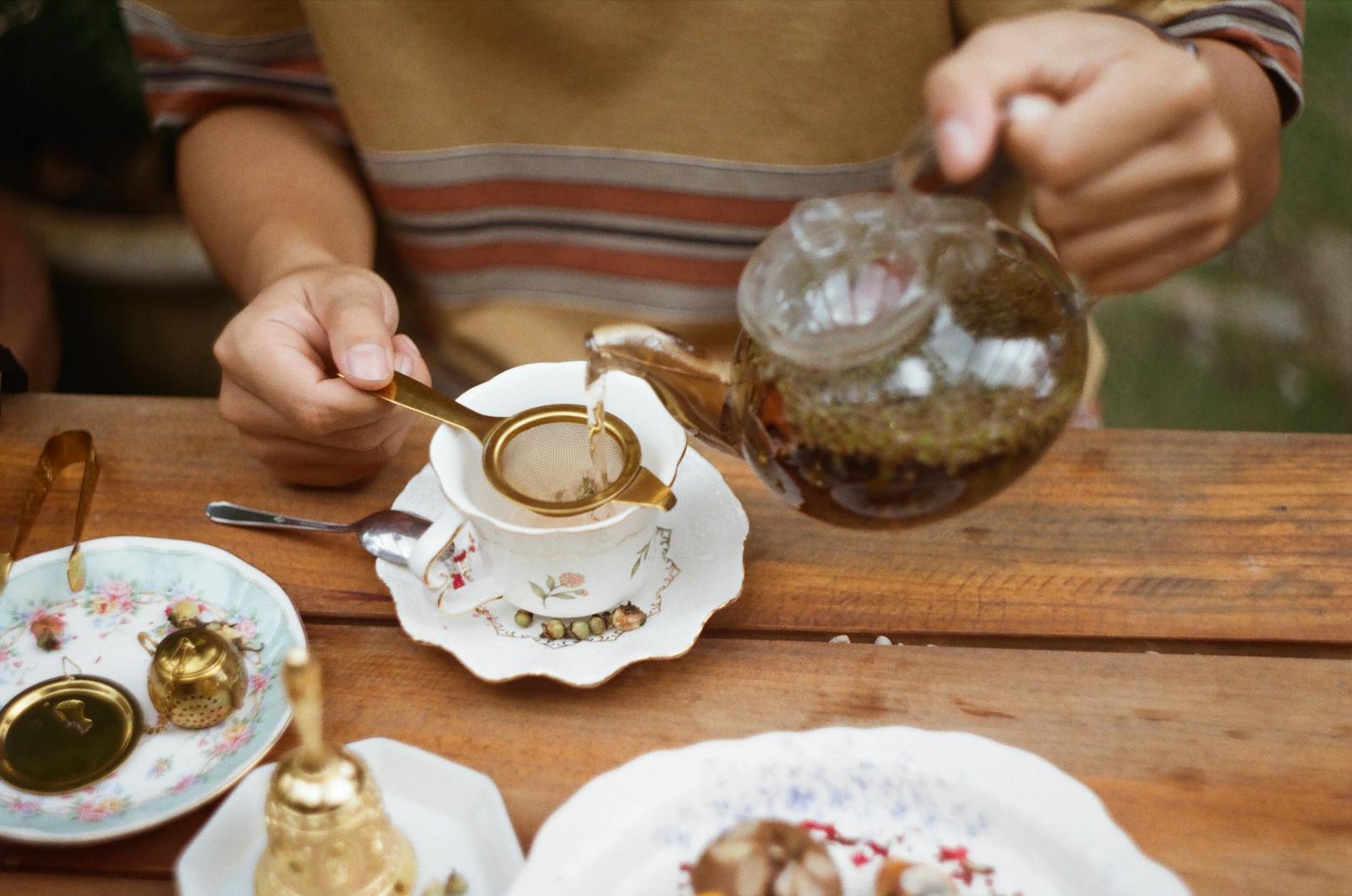 Close-up of a person pouring tea into a porcelain cup with floral design on a wooden table.