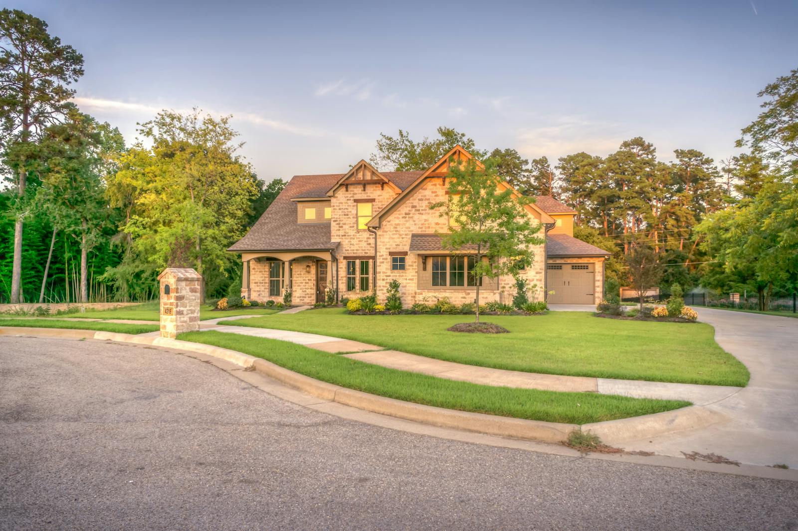 Elegant stone family home with manicured lawn, trees, and driveway on a sunny day.