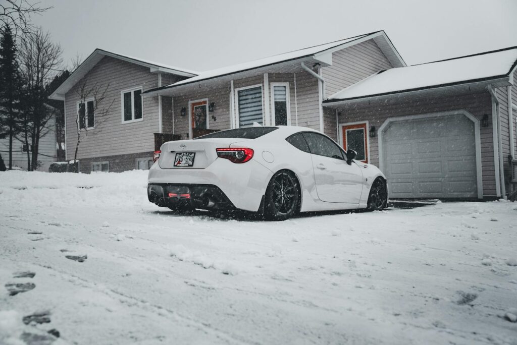A white sports car parked in a snowy suburban driveway during winter snowfall.
