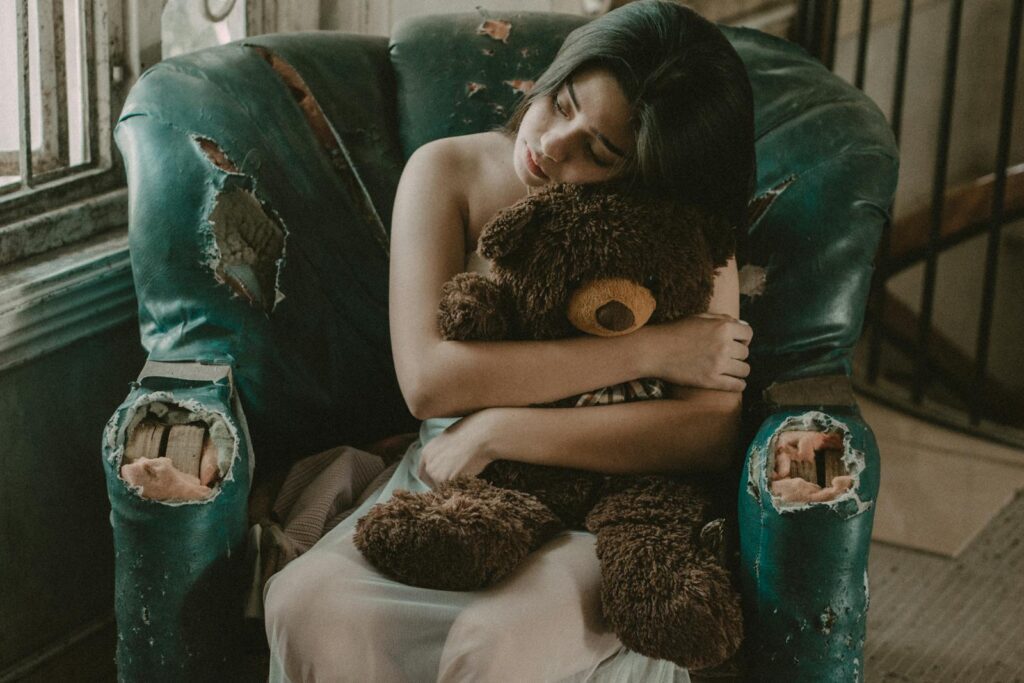 A young woman in a vintage chair embraces a teddy bear, evoking a sense of nostalgia and comfort.