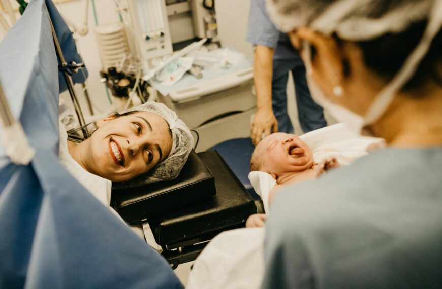 A mother smiles lovingly at her newborn in a hospital delivery room, attended by medical staff.