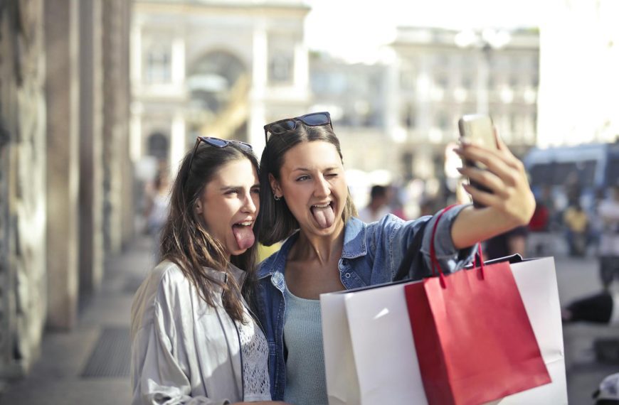 Two young women taking a playful selfie while shopping outdoors in an urban setting.