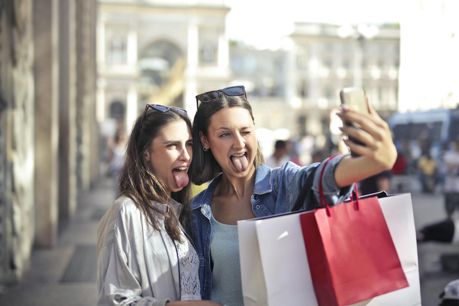 Two young women taking a playful selfie while shopping outdoors in an urban setting.