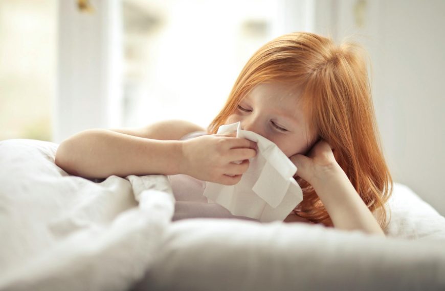 Redheaded child resting in bed, wiping nose with tissue, unwell with flu-like symptoms.