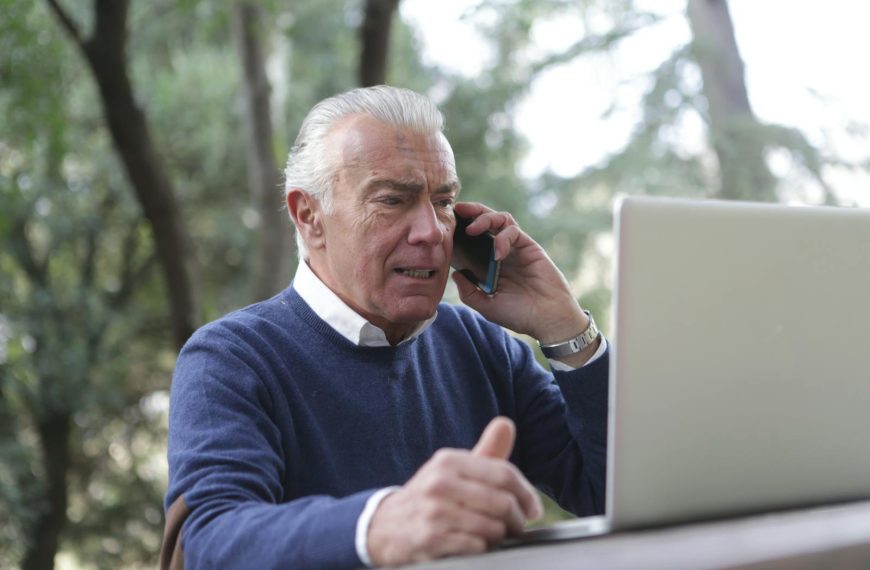 Elderly man using laptop and smartphone outdoors, showcasing modern connectivity.