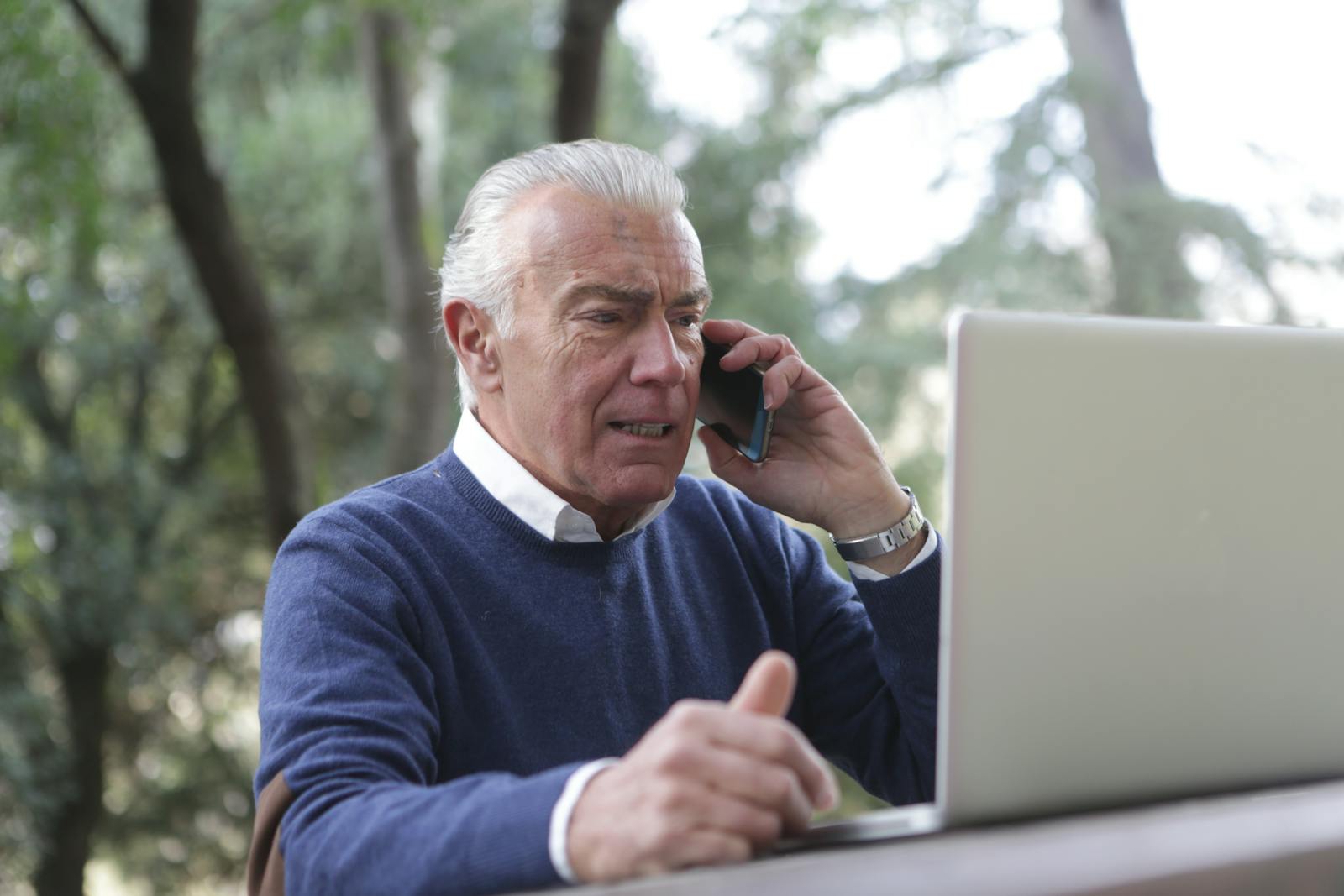 Elderly man using laptop and smartphone outdoors, showcasing modern connectivity.