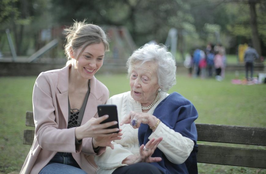 Delighted female relatives sitting together on wooden bench in park and browsing mobile phone while learning using