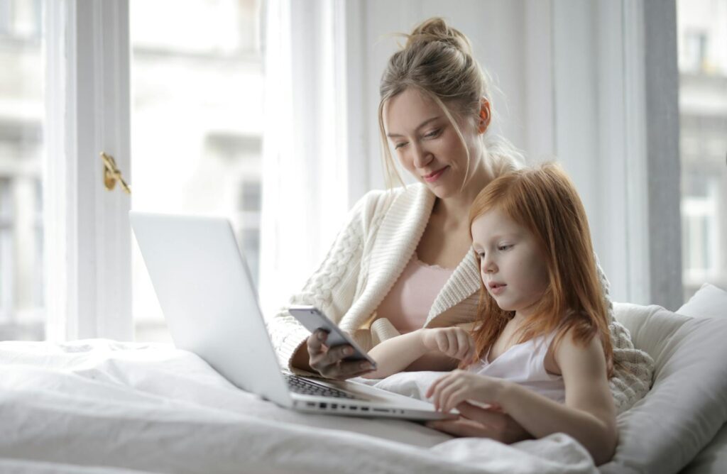 A mother and daughter enjoying time together with a laptop and phone in a cozy bedroom setting.