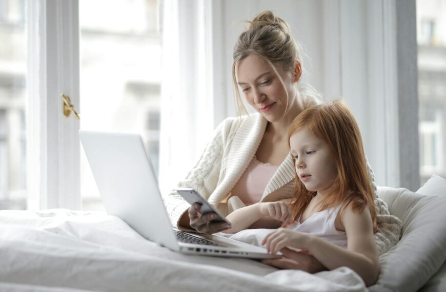 A mother and daughter enjoying time together with a laptop and phone in a cozy bedroom setting.