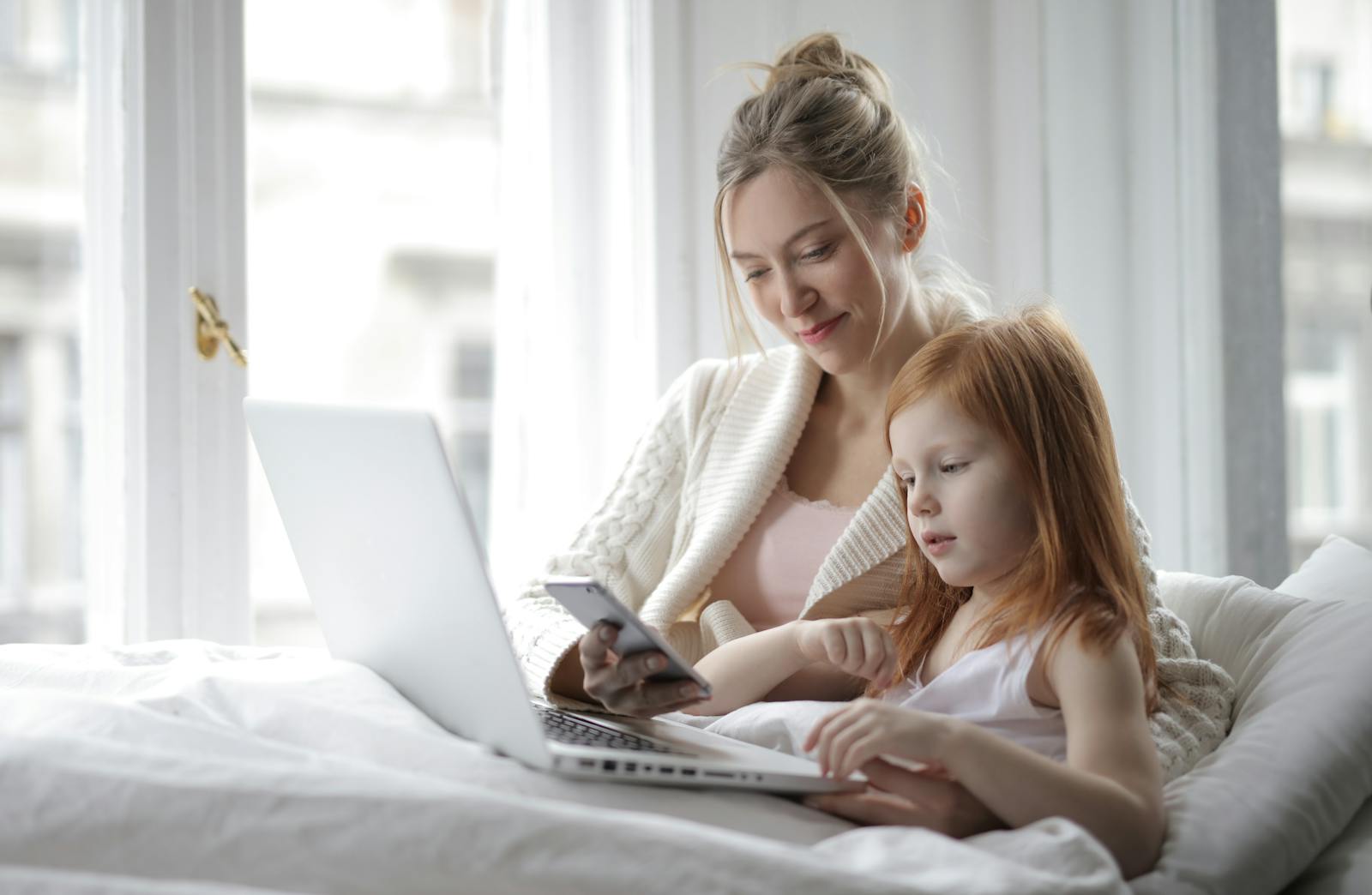 A mother and daughter enjoying time together with a laptop and phone in a cozy bedroom setting.