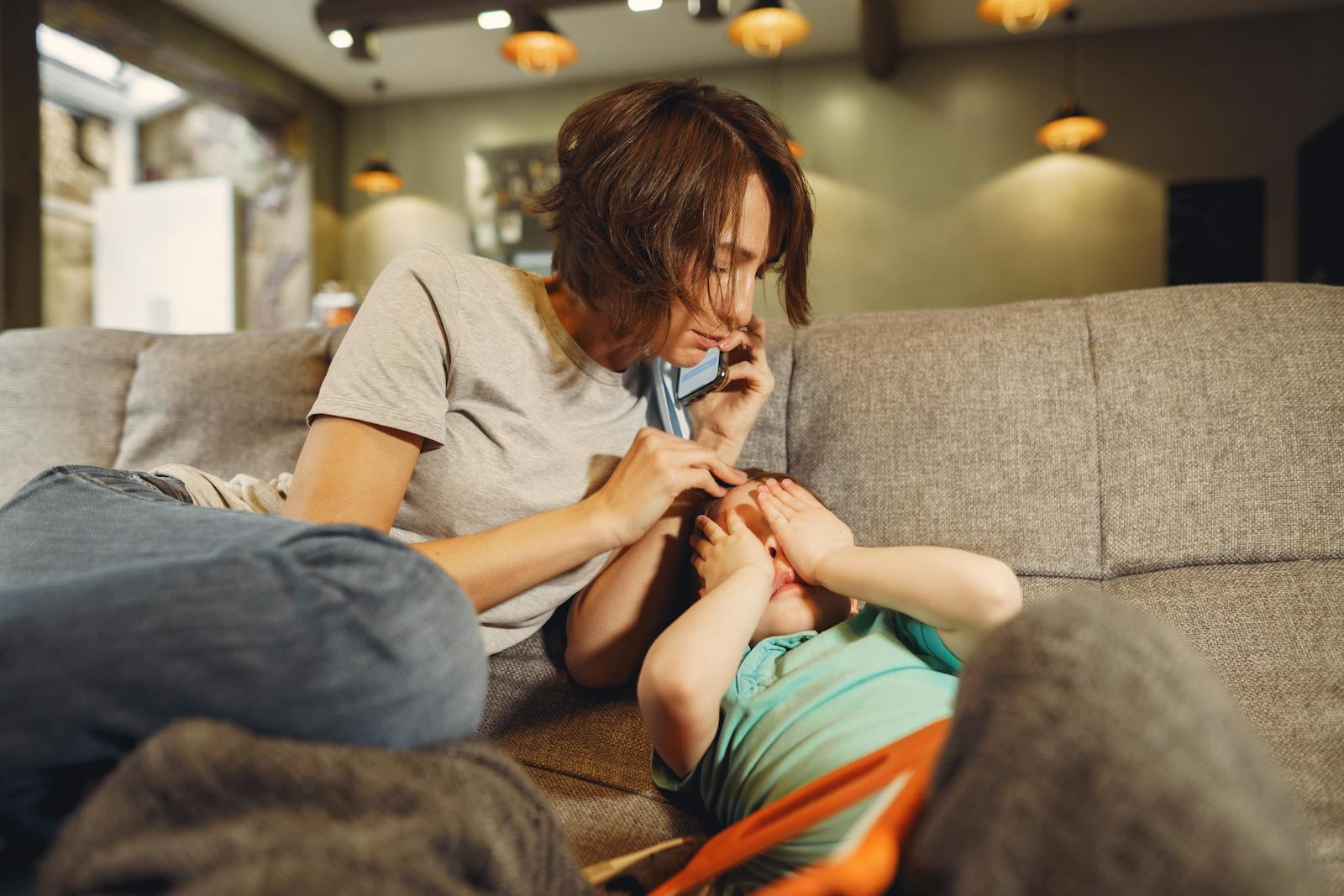 A caring mother on the phone comforts her child on the sofa in a cozy living room.
