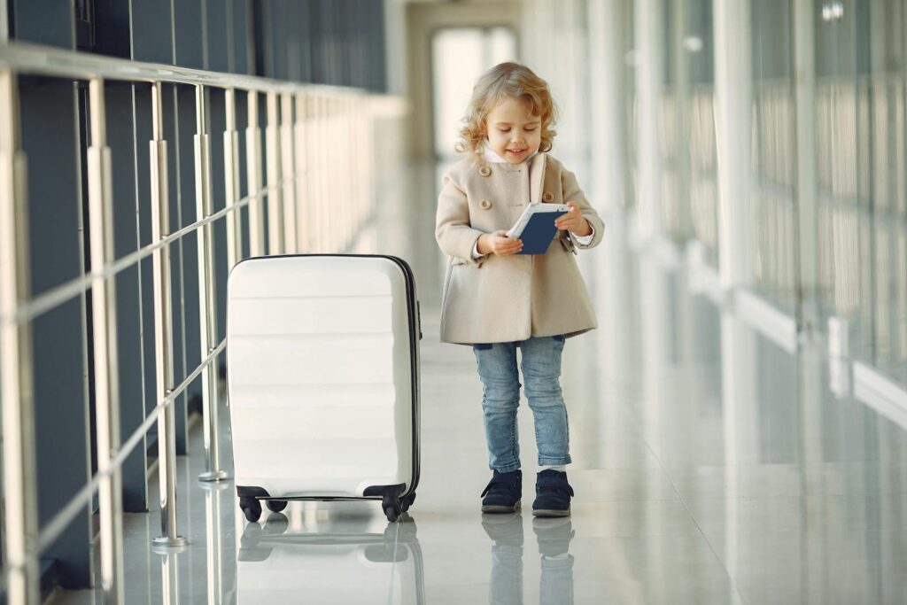 Full body of smiling cute little girl in jeans and beige coat standing near suitcase and checking information in documents