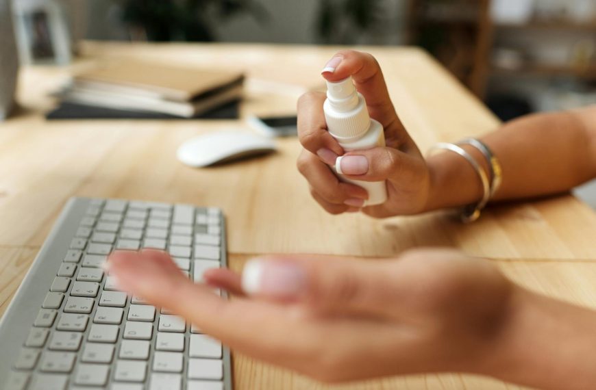 Person sanitizing hands at a desk with spray bottle, keyboard, and computer mouse.