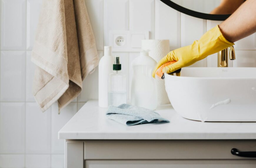 Person cleaning a bathroom sink with yellow gloves and cleaning products on a countertop.