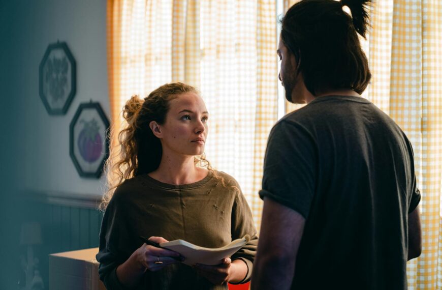 Unemotional focused young couple wearing casual outfits standing near window with notebook and looking at each other seriously