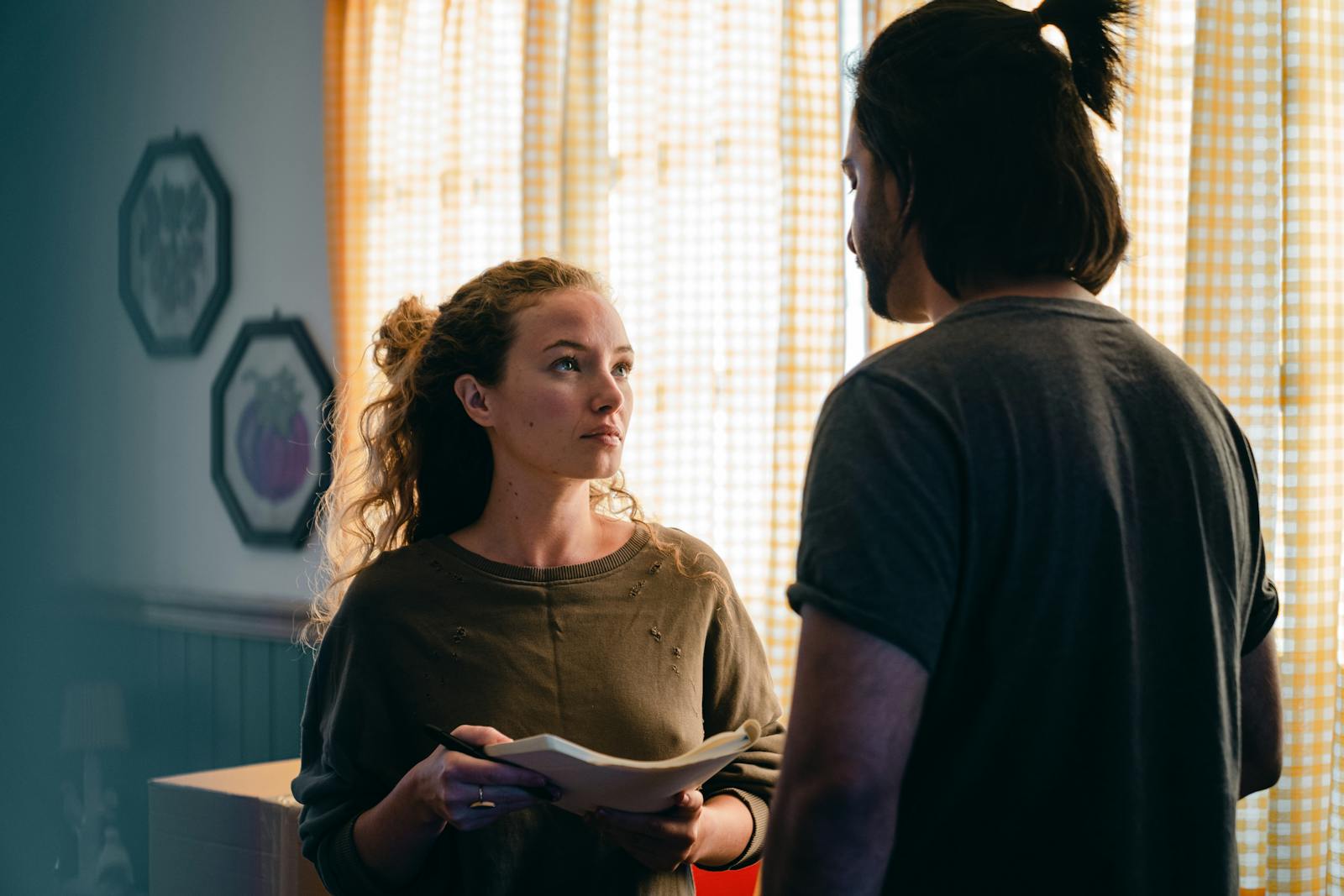 Unemotional focused young couple wearing casual outfits standing near window with notebook and looking at each other seriously