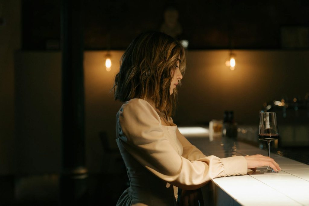 A stylish woman in a soft dress sits at a bar, subtly lit with warm lighting, holding a glass of wine.